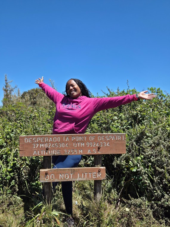 Diana outdoors on a hillside with arms outstretched by a trail marker.